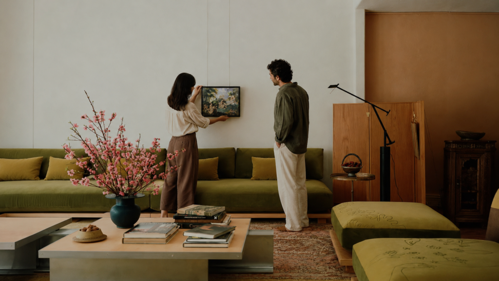 A couple installs a small framed painting in a stylish living room with a green sofa, showing how art collecting can begin with interiors but should grow beyond matching the couch.